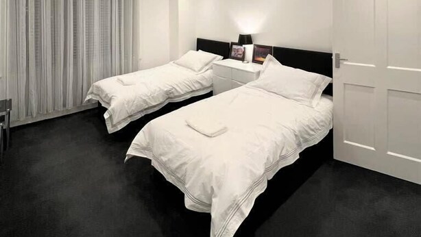 Una habitación de hotel moderna con dos camas individuales, una mesita de noche con lámpara, un ventanal con cortinas y una puerta.