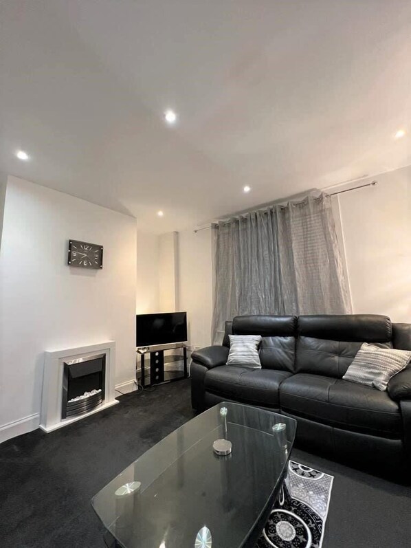 A modern living room with a black leather sofa, a glass coffee table, a flat-screen TV, and a fireplace.