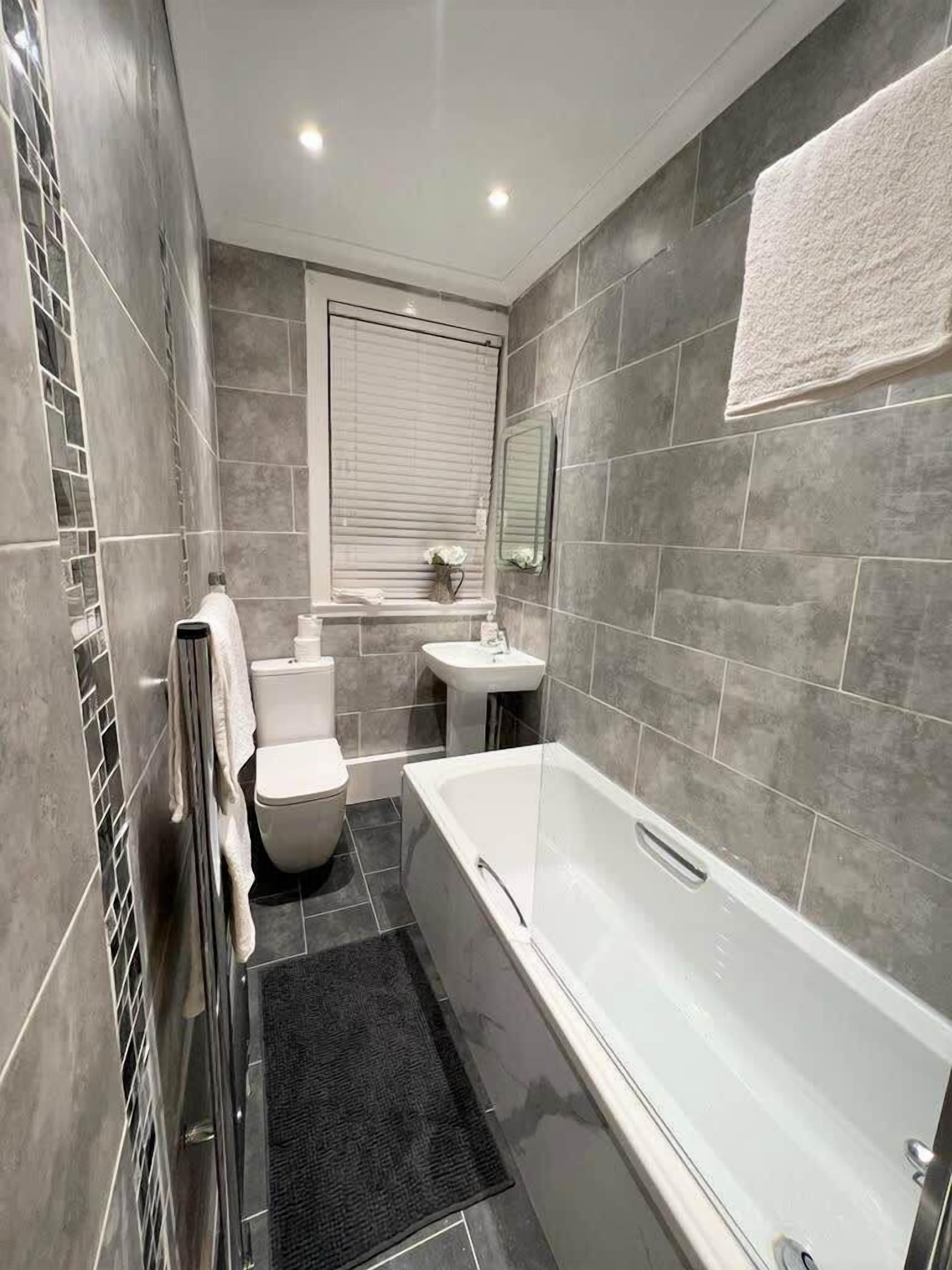 A modern bathroom with a bathtub, toilet, and sink, featuring grey tiles and a window with blinds.