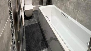 Un baño moderno con bañera, inodoro y lavamanos, con baldosas grises y una ventana con persianas.