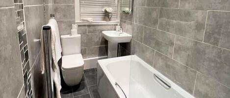 A modern bathroom with a bathtub, toilet, and sink, featuring grey tiles and a window with blinds.