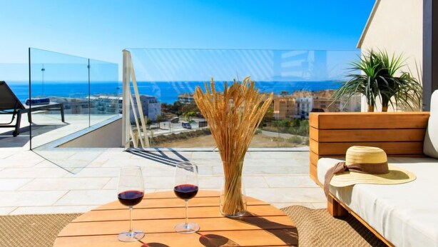 A balcony with a glass railing, a wooden table with two wine glasses and a vase, a sofa, and a view of the sea and buildings. - Marinsa Beach II Sea View/Roof terrace by Interhome (Torrox Costa)