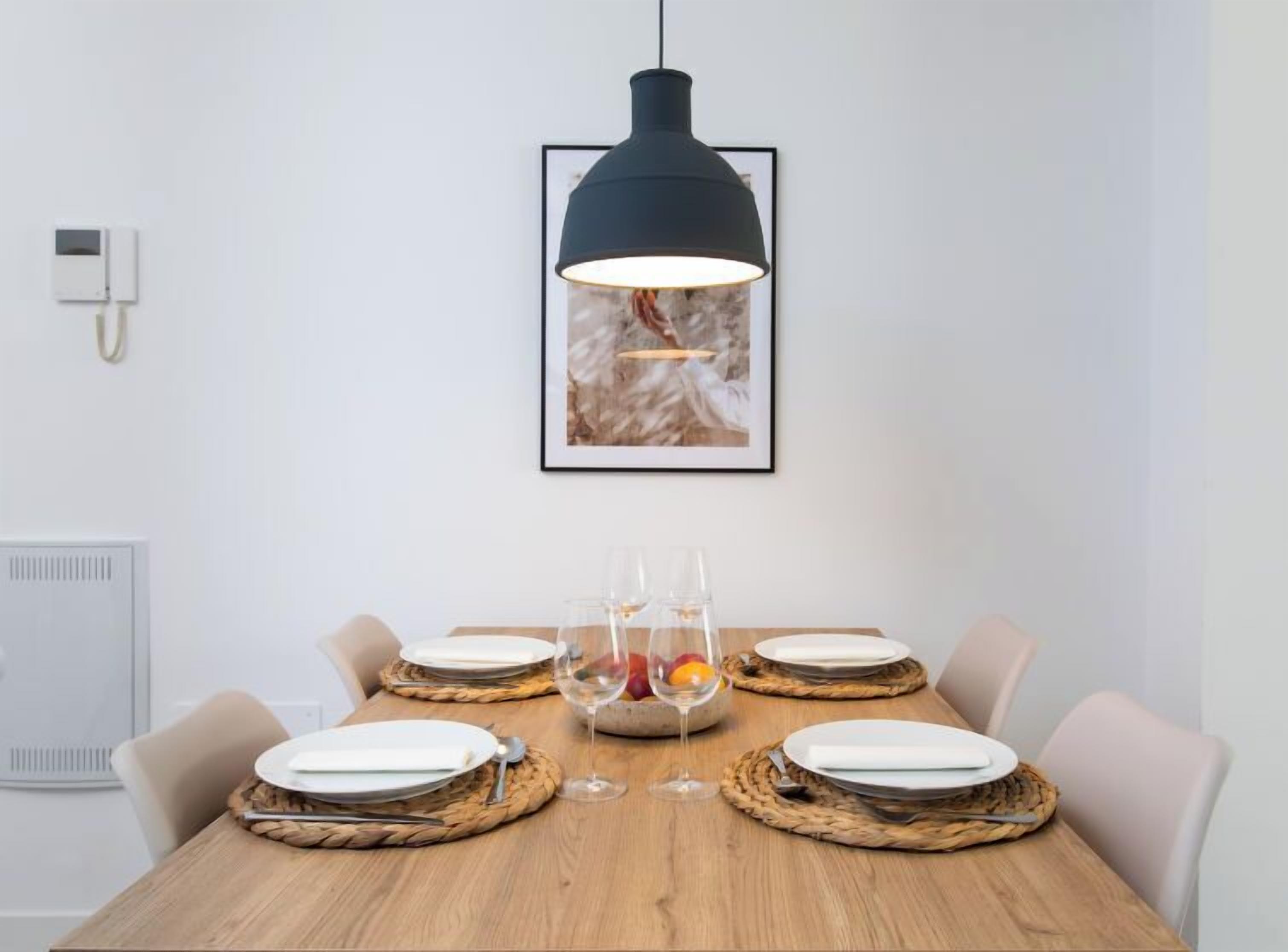 Une table à manger dressée avec des assiettes, des couverts et un verre de vin, un luminaire suspendu au-dessus et un tableau encadré au mur.