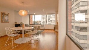 A modern living room with a round dining table, white wireframe chairs, a hanging light fixture, and a large window offering a view of high-rise buildings.