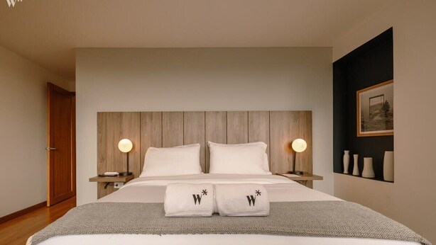 A modern hotel room with a large bed, two bedside lamps, a wooden headboard, and a framed picture on the wall.