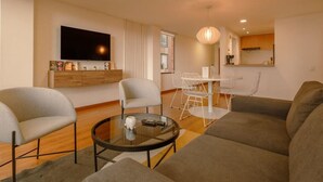 A modern living room with a large flat-screen TV, a sofa, two armchairs, a glass coffee table, and a dining area with a white table and chairs.
