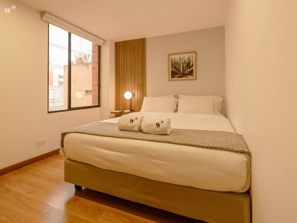 Une chambre à coucher avec un grand lit, une table de chevet avec une lampe, une fenêtre et une œuvre d’art accrochée au mur.