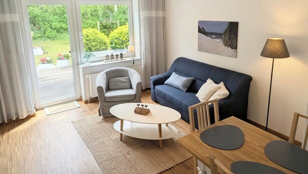 A modern living room with a sofa, armchair, coffee table, and dining table. - Apartment with private wooden terrace and beach chair (Friedrichskoog)