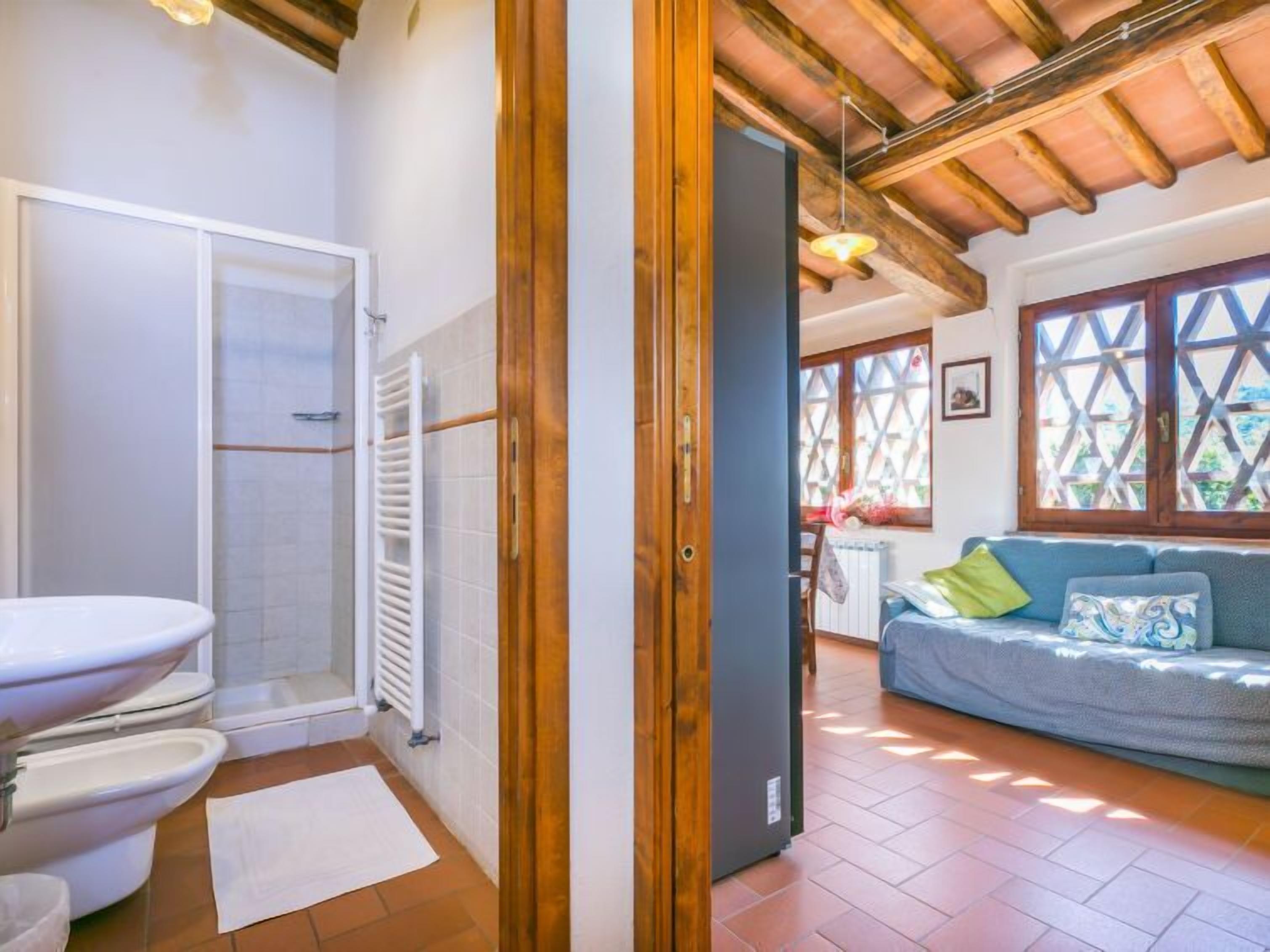 Ein Badezimmer mit Toilette, Dusche und Heizkörper. Eine Holztür führt in einen Wohnbereich mit Sofa und Fenster.