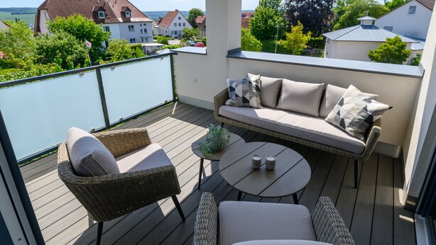 Ein Balkon mit einer Rattan-Couch, zwei Stühlen, einem kleinen Tisch und einem Glasgeländer.