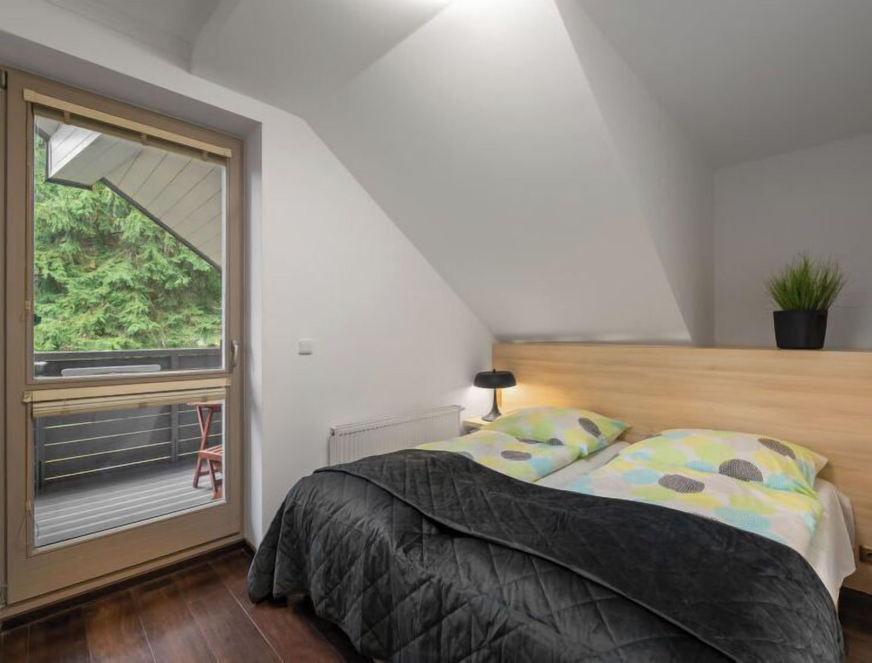 A bedroom with a bed, a window with a view of greenery, a radiator, a nightstand with a lamp, and a small plant on the wall.