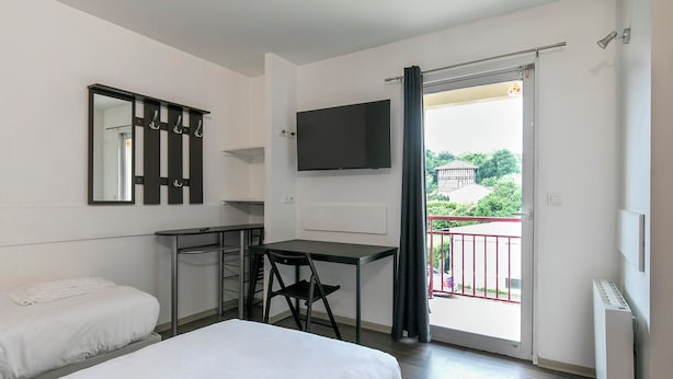 A bedroom with a bed, a desk, a chair, a TV, and a balcony with a view of greenery and a building. - Apparts Confort 87 (Limoges)