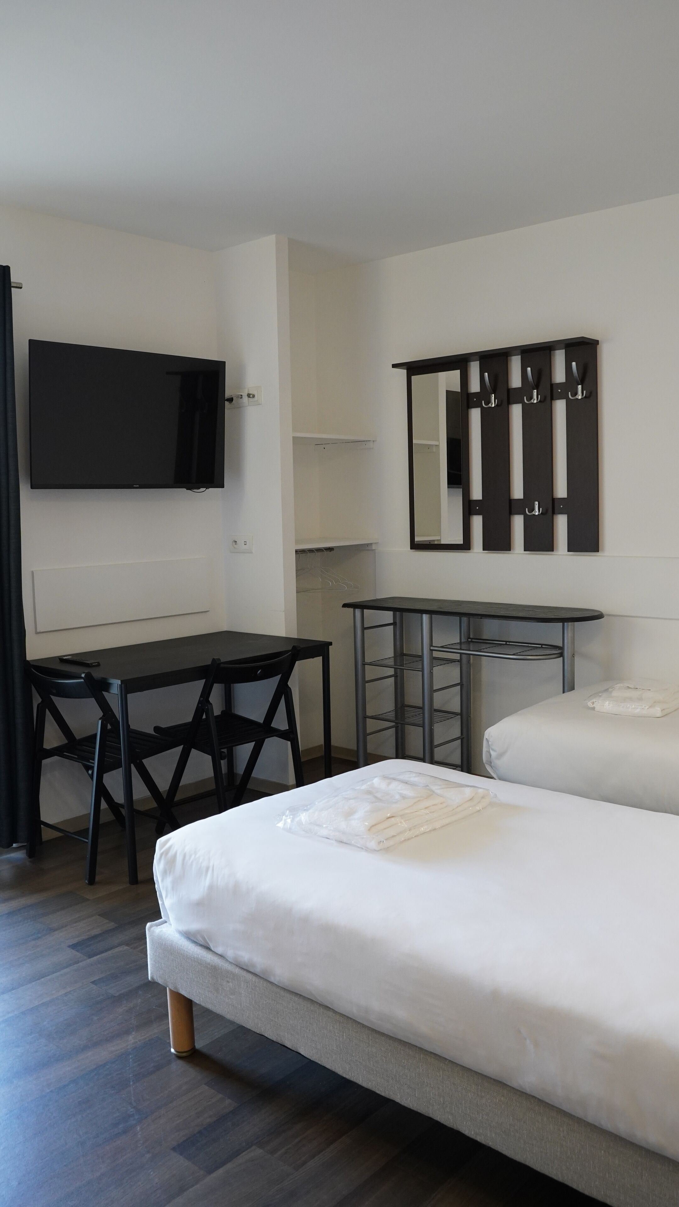 Quarto de hotel com cama, escrivaninha, cadeira, televisão, espelho e um cabideiro.