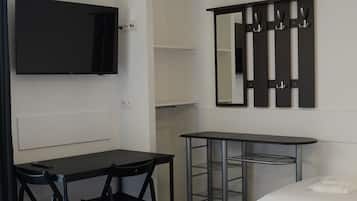 Quarto de hotel com cama, escrivaninha, cadeira, televisão, espelho e um cabideiro.