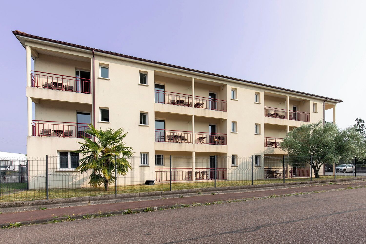 Un edificio residencial de varios pisos con balcones, un patio cercado y una calle en la parte delantera.