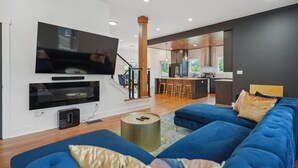 A modern living room with a large flat-screen TV, a blue sofa, and a kitchen area in the background. - New Modern 4BR Smart Home Downtown Atlanta GWCC (Atlanta)