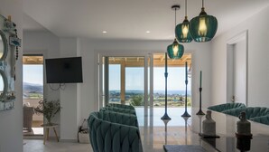 A modern living room with a view of the ocean, teal upholstered chairs, and pendant lighting. - Villa Bella, Mikri Vigla.  Private pool, jacuzzi & amazing views. Sleeps 10+. (Naxos)