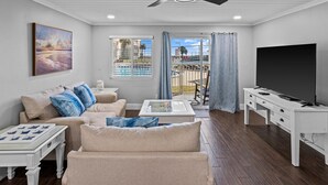 Un salon moderne doté d’un ventilateur de plafond, d’un ensemble de canapés, d’une table basse, d’un téléviseur et offrant une vue sur une piscine par une fenêtre.