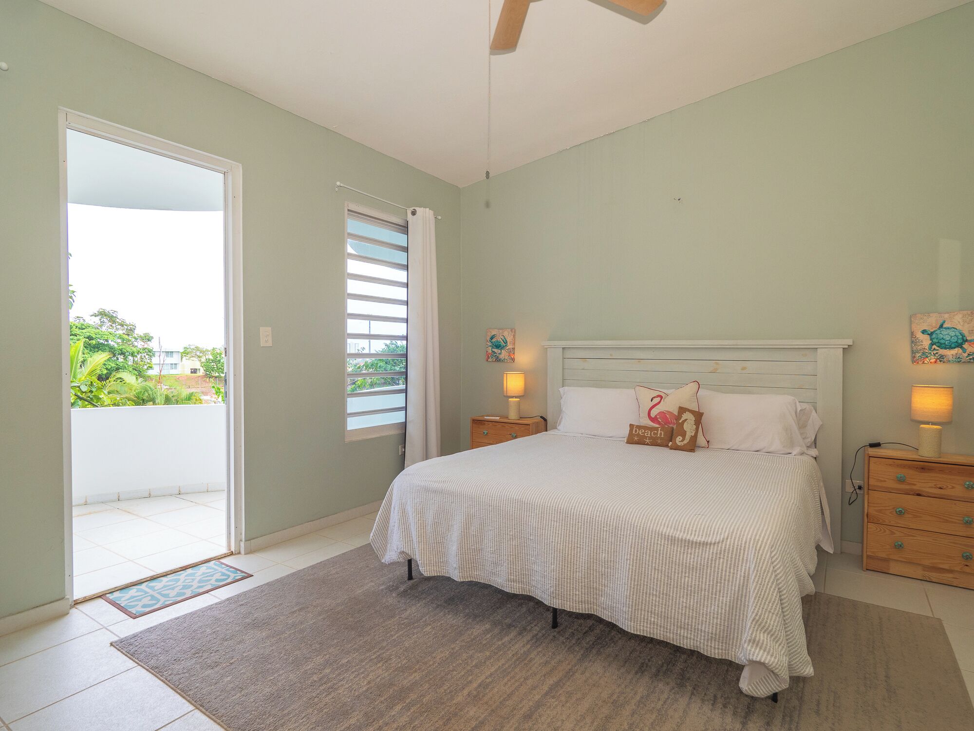A bedroom with a bed, bedside tables, a window with blinds, and a view of greenery outside.