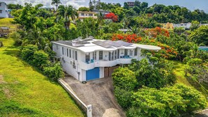 A white building with solar panels, surrounded by lush greenery and a clear sky. - Hideaway with Wraparound Balcony: Beautiful Views, Great Vibes, Family Friendly (Luquillo)