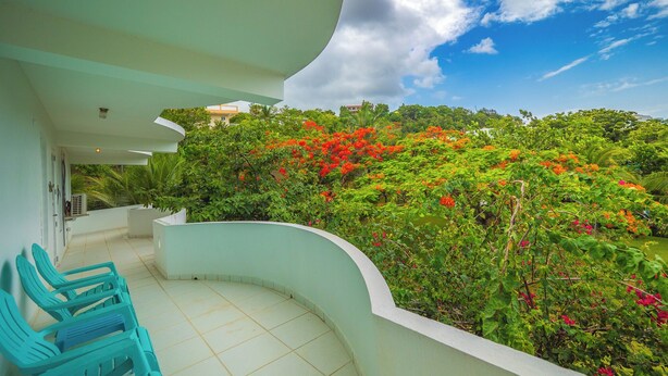A balcony with a curved white railing, a tiled floor, and a view of lush greenery and red flowers. - Hideaway with Wraparound Balcony: Beautiful Views, Great Vibes, Family Friendly (Luquillo)