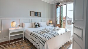 Une chambre d’hôtel avec un lit bien fait, une table de chevet avec une lampe, une petite plante en pot et une fenêtre donnant sur des immeubles.