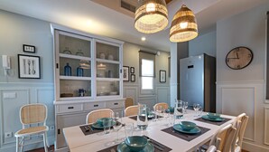 Un coin repas avec une longue table dressée pour quatre personnes, entourée de chaises en osier. On y trouve deux lustres suspendus en osier, un buffet blanc avec des portes en verre et une horloge murale.