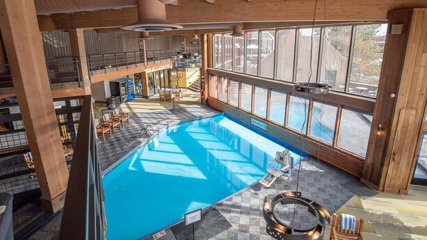 A large indoor swimming pool with a view of the outside, wooden architecture, and a seating area with chairs.
