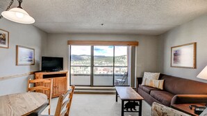 A living room with a wooden dining table, chairs, a sofa, a television, and a balcony with a view of the city.