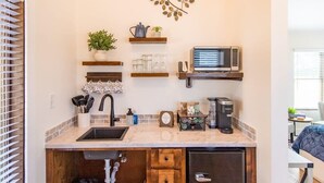 Cocina compacta con lavabo, microondas y cafetera. Hay repisas sobre la encimera con utensilios de cocina y un adorno de pared.