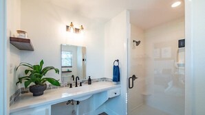 Un baño con tocador blanco, espejo, planta en maceta y zona de ducha.