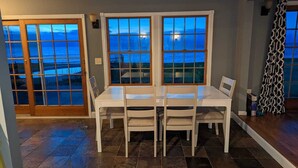Un coin repas avec une table et des chaises, de grandes fenêtres donnant sur l’océan et un rideau à motifs sur le côté droit.