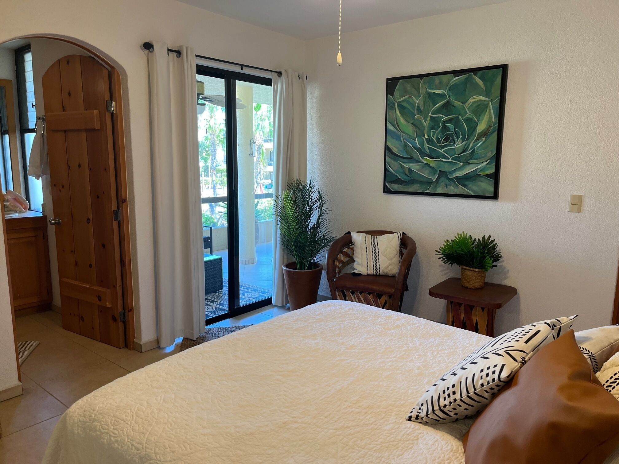 A bedroom with a large bed, a chair, a small table with a plant, and a door leading to a balcony.