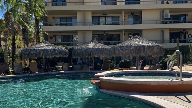 Una piscina con agua cristalina, rodeada de tumbonas y sombrillas, con un edificio de hotel al fondo.