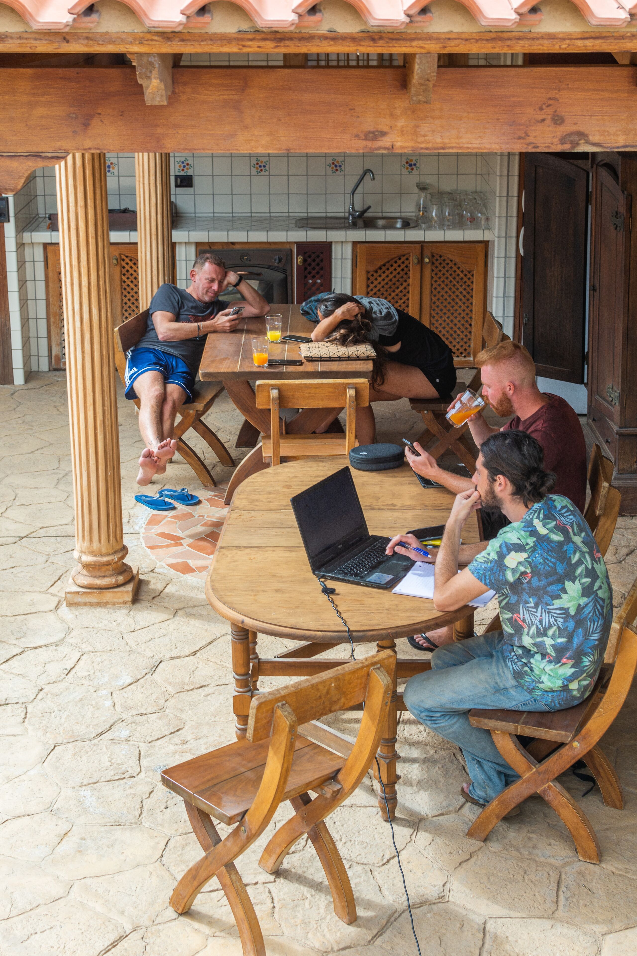 Un groupe de personnes est assis à des tables en bois, à l’abri sous un toit en tuiles, et s’adonne à diverses activités, comme utiliser des ordinateurs portables ou boire des boissons.