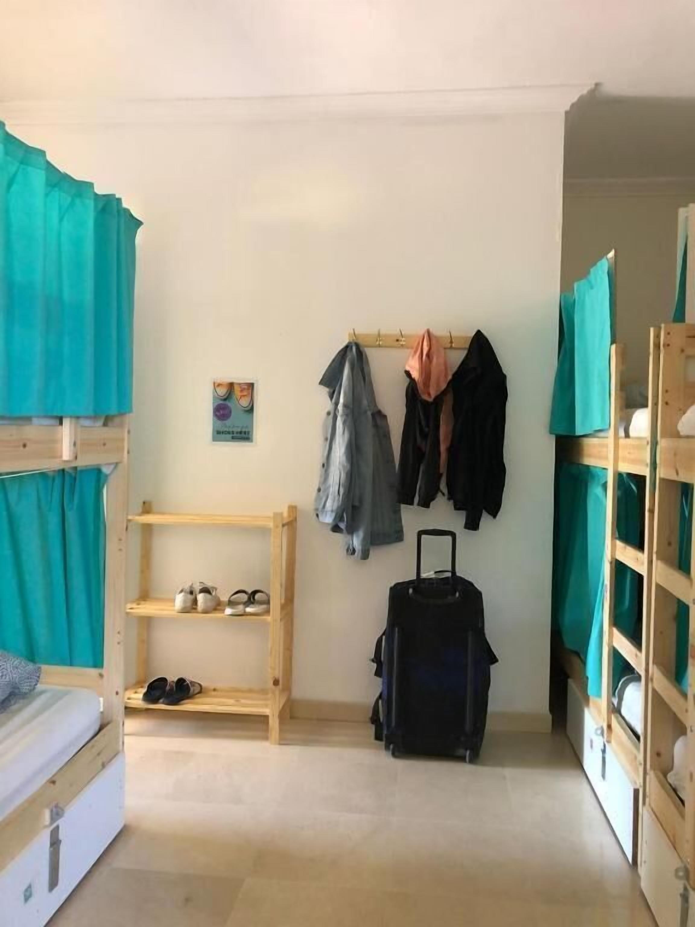 A dormitory room with bunk beds, a small wooden shelf with shoes, a wall-mounted coat rack, and a suitcase.