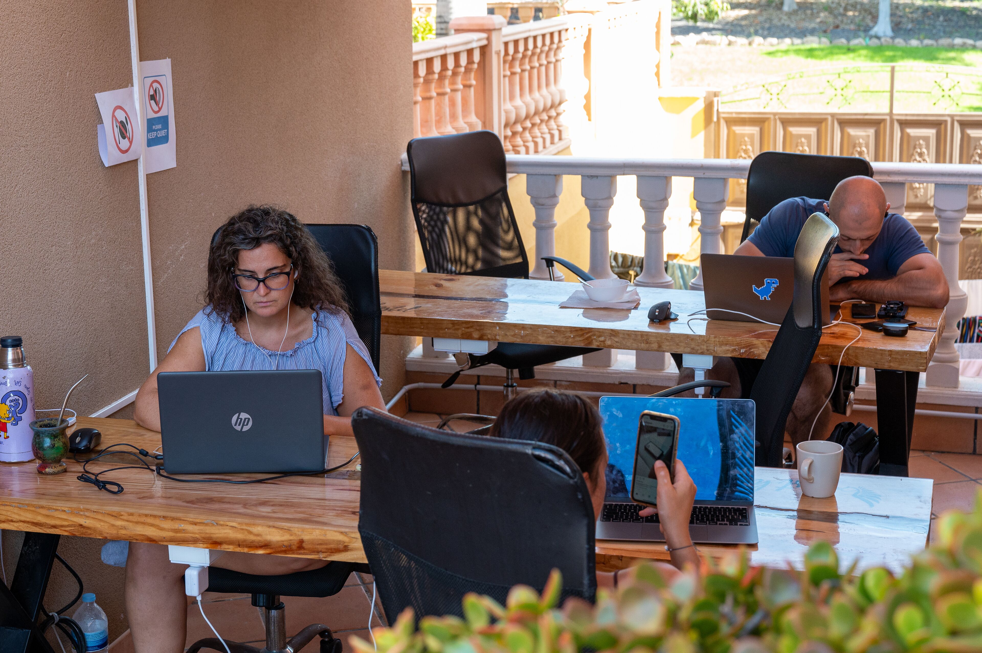 Een groep mensen werkt aan buitentafels met laptops, omringd door stoelen en een hekwerk.