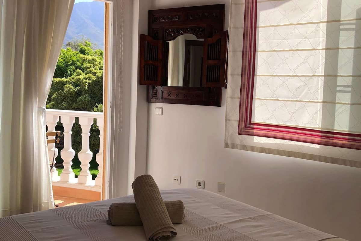 A bedroom with a bed, a window with curtains, a mirror, and a view of greenery outside.
