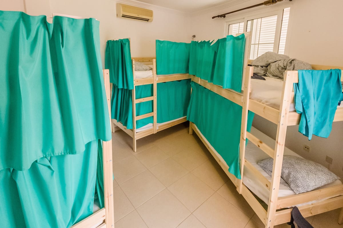 A dormitory room with bunk beds, teal curtains, a ceiling light, and a window with blinds.