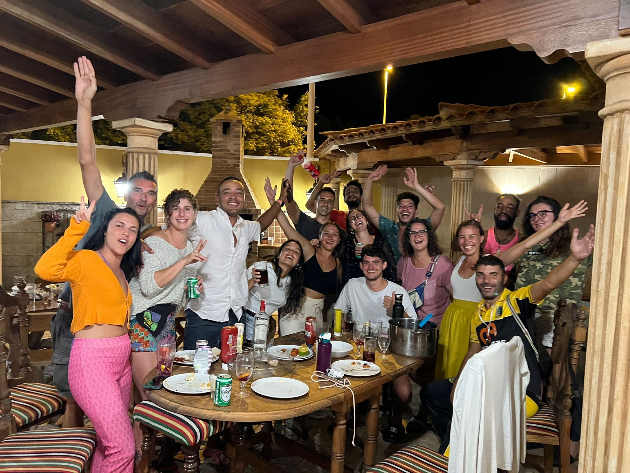 Un grupo de personas sentadas a una mesa con diversos alimentos y bebidas, rodeadas de muebles y pilares de madera.