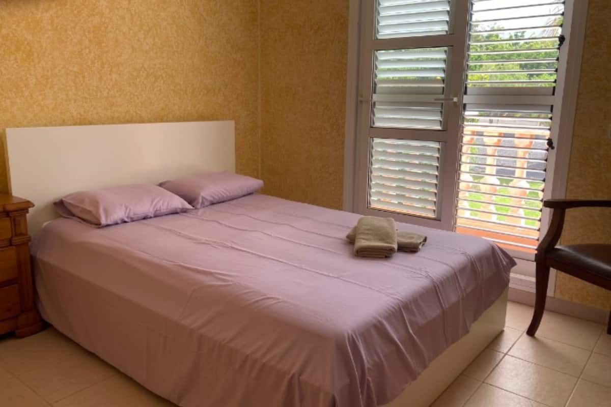 A bedroom with a bed, pillows, a nightstand, a chair, and a window with blinds.