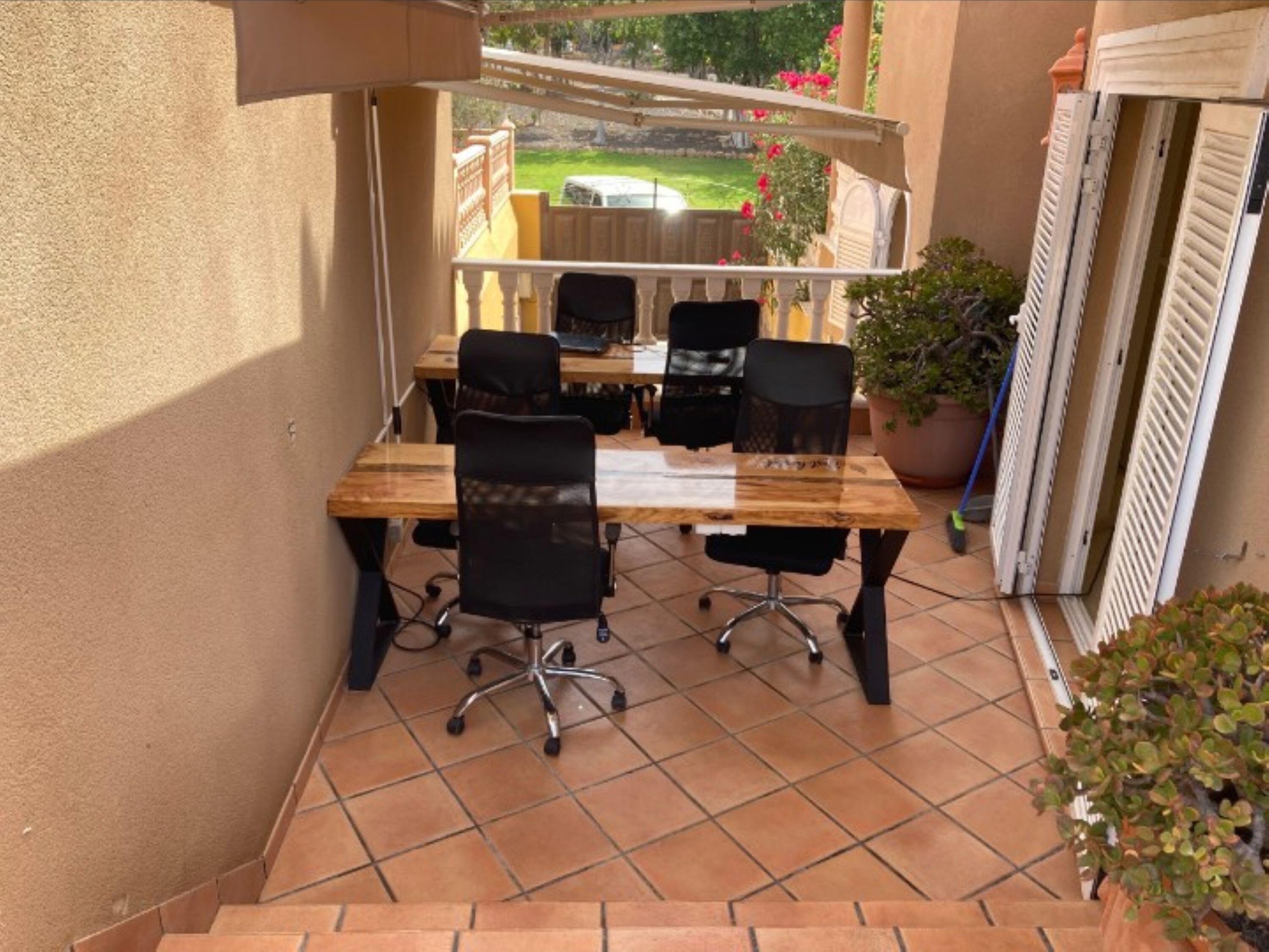 A patio with a wooden table and four black office chairs, surrounded by beige walls and a tiled floor.