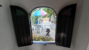 A view through a window with open shutters, revealing outdoor seating with plastic chairs and a table, a colorful umbrella, and marigold flowers. - Pousada Toa Toa Em Pipa (Tibau do Sul)