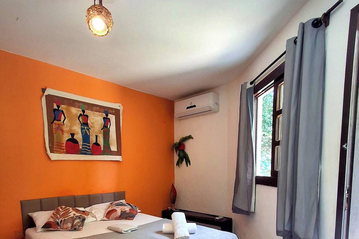 A hotel room with a bed, a painting on the wall, an air conditioning unit, and a window with curtains.