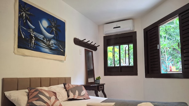 A bedroom with a bed, pillows, a blanket, an air conditioning unit, a window with shutters, and a painting on the wall. - Pousada Toa Toa Em Pipa (Tibau do Sul)