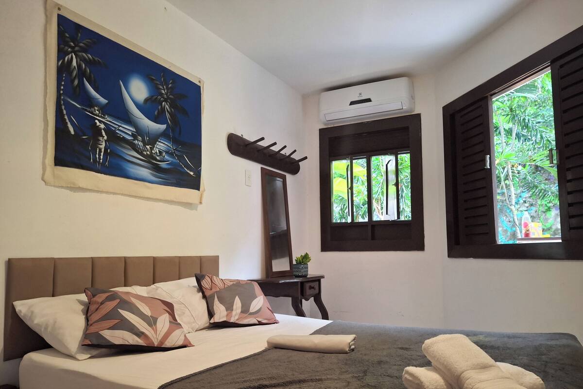 A bedroom with a bed, pillows, a blanket, an air conditioning unit, a window with shutters, and a painting on the wall.