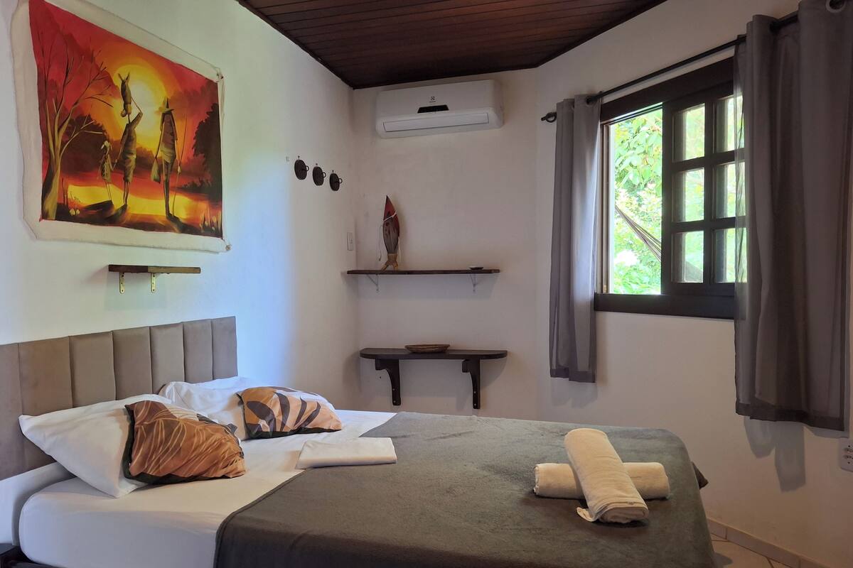 A room with a bed, a painting, an air conditioner, a window with curtains, and a small table.