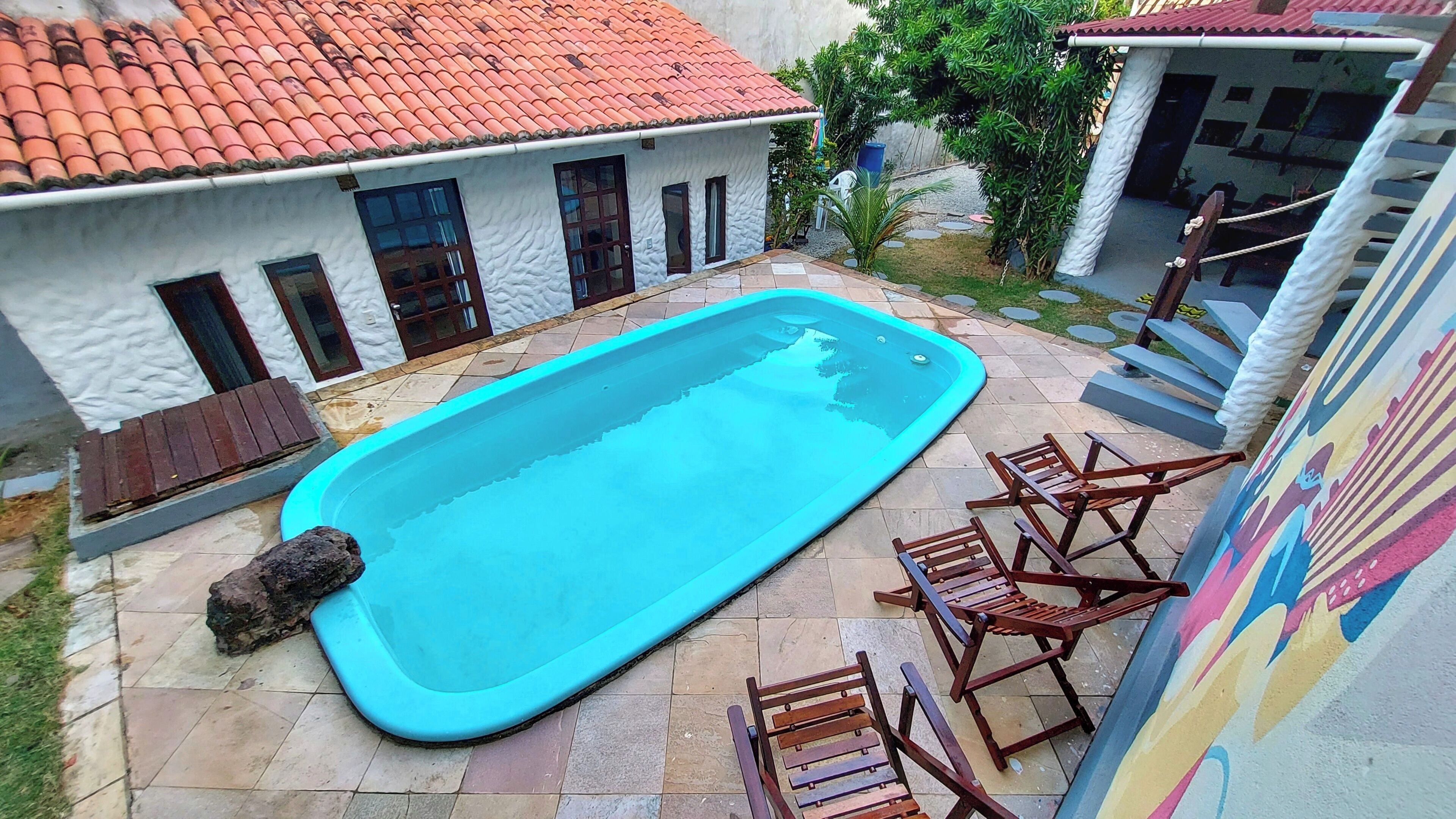 A swimming pool with a tiled deck, wooden chairs, and a white building with a red-tiled roof.