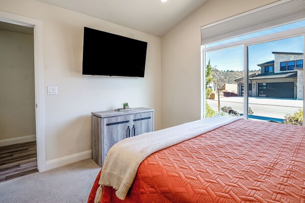A bedroom with a large bed, a TV mounted on the wall, a small cabinet, and a sliding glass door leading to a garage.