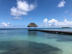 Un cielo azul despejado, un muelle de madera que conduce a una estructura de techo de paja sobre aguas turquesas tranquilas.